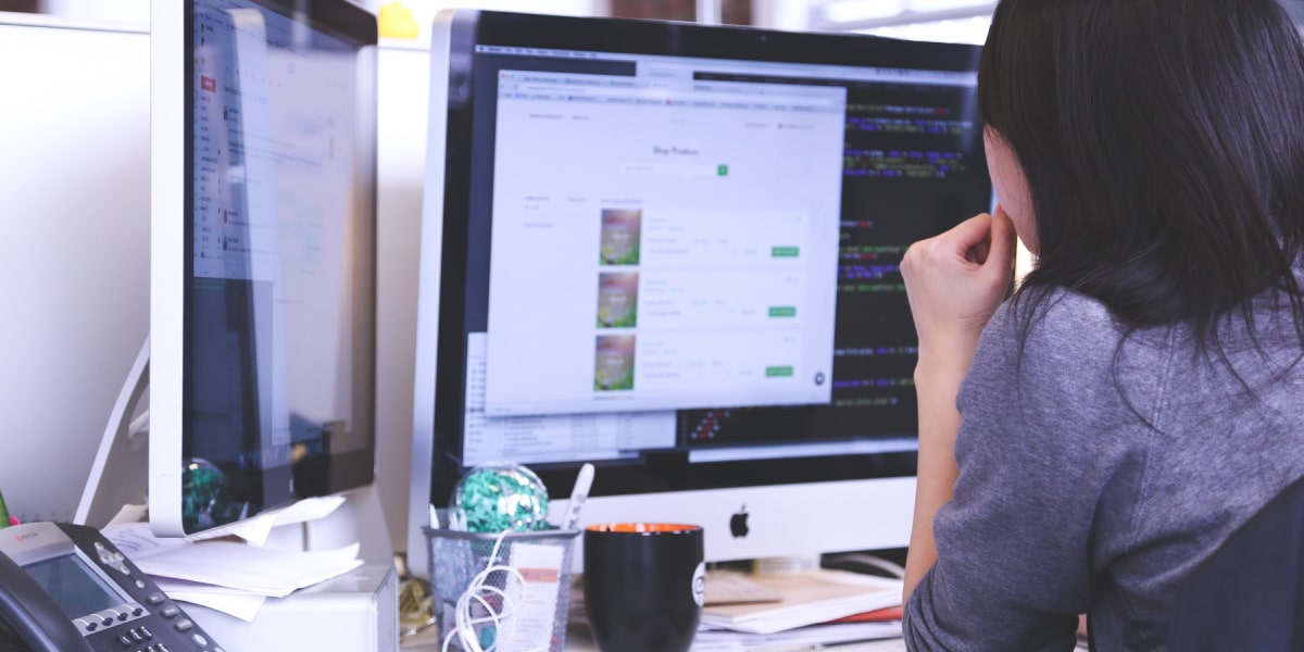 A web developer sits at their workstation, looking at HTML code and markup on two screens.