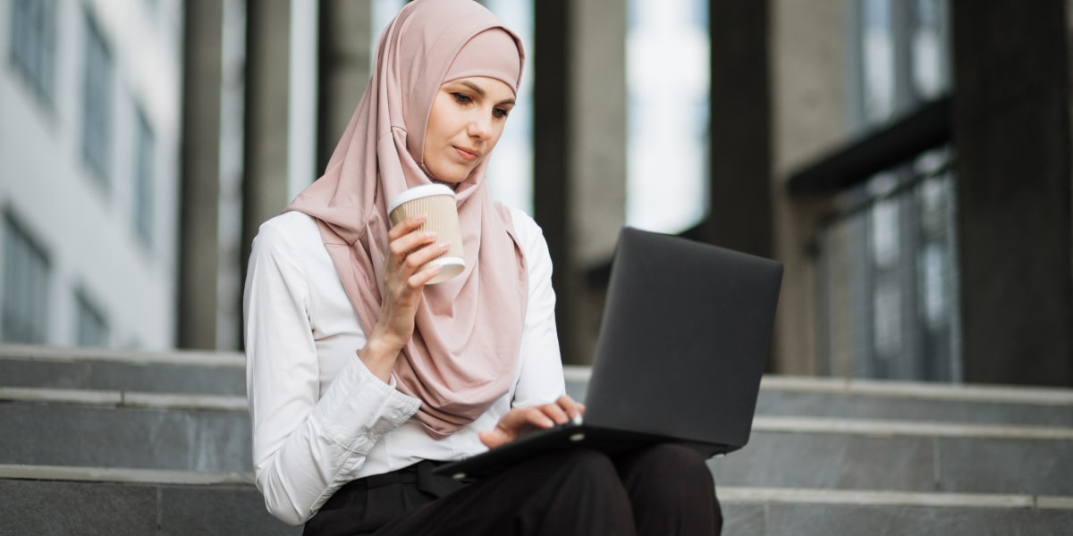 A digital marketing degree student sitting on a step outside, holding a cup of coffee and looking at a laptop