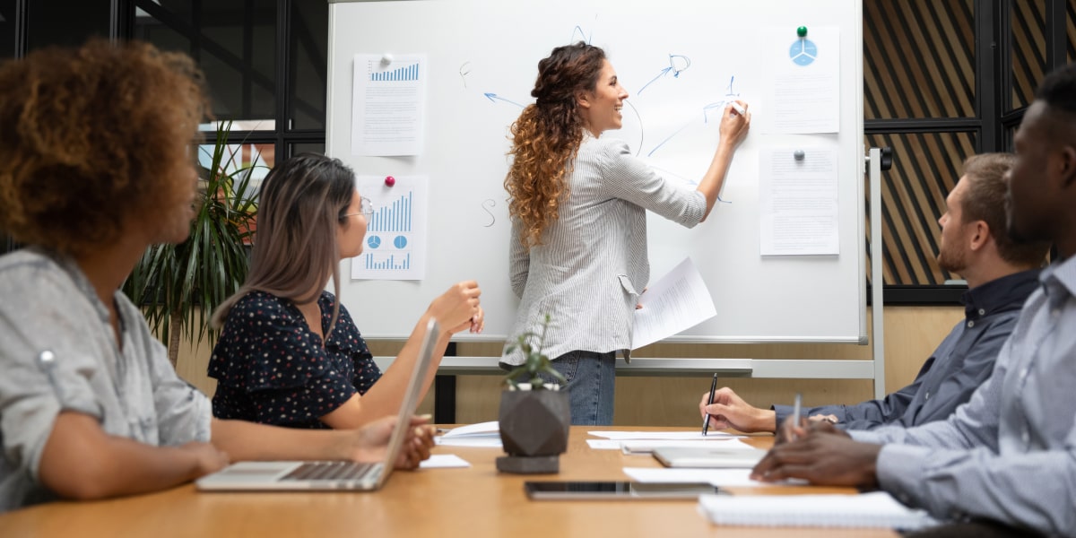 A group of digital marketing degree students sitting around a desk, watching a lecturer draw on a whiteboard