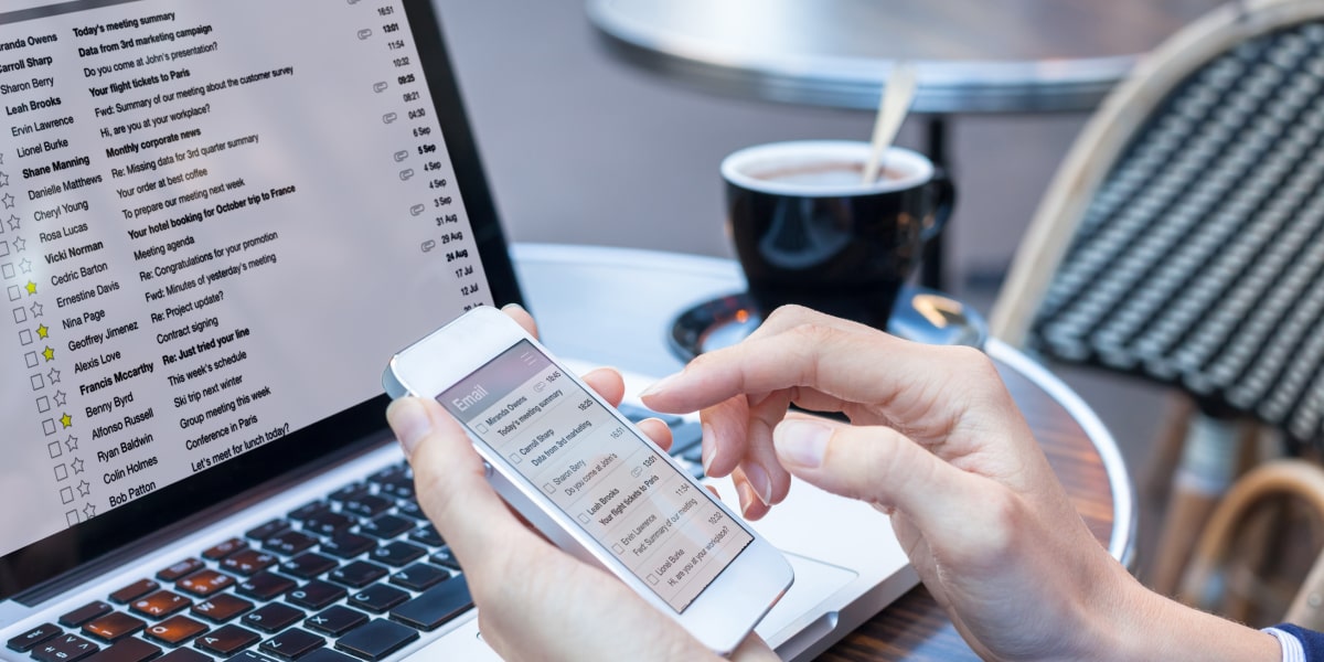 A person reading emails on a laptop and a smartphone with a cup of coffee in the background