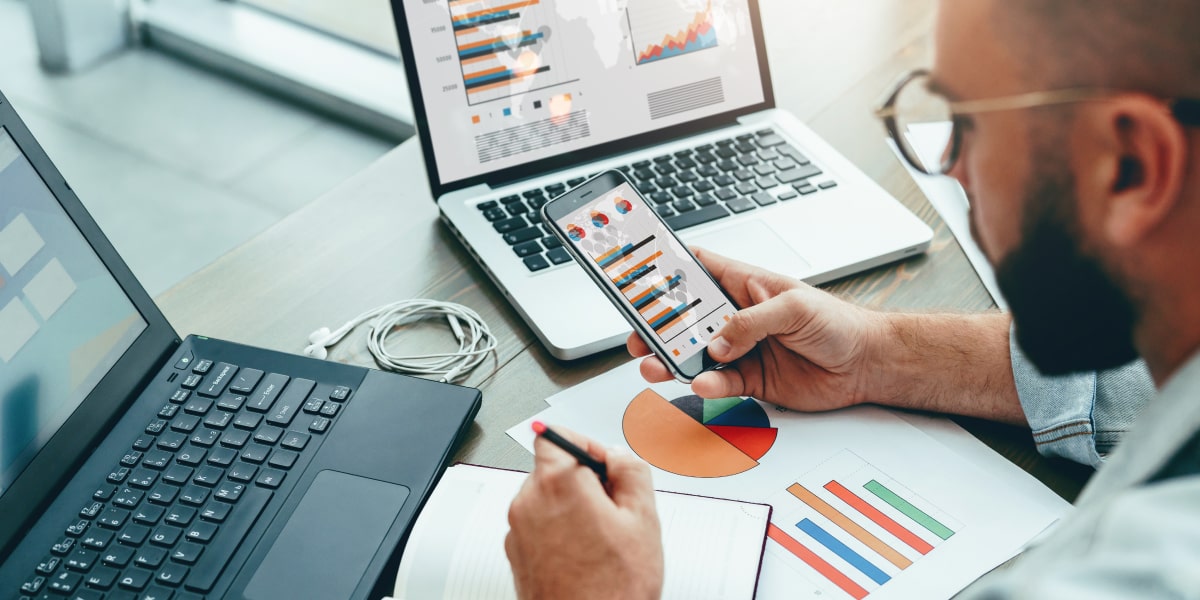 A marketing professional holding a smartphone and looking at graphs on a laptop screen