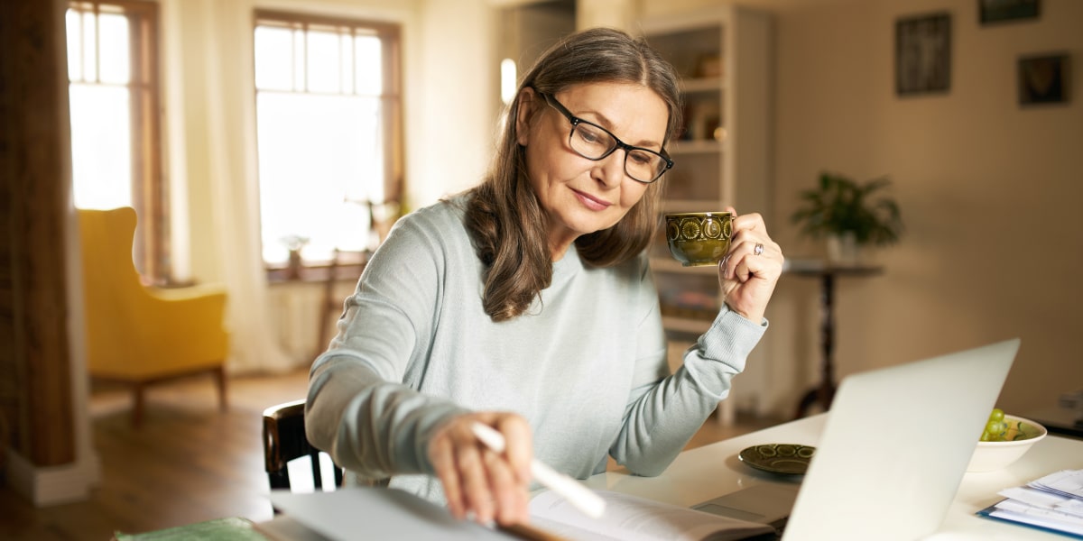 Image of an older female web developer works from home at her laptop holding a mug of tea.