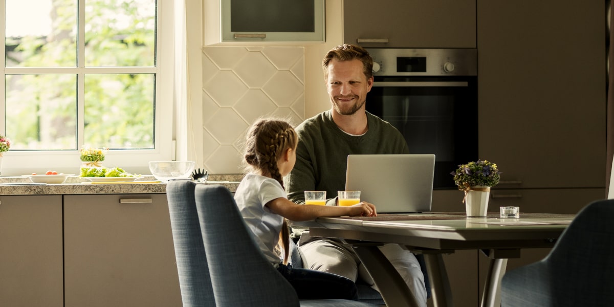 A web developer sits at his kitchen table with a laptop open and his daughter sitting beside him.
