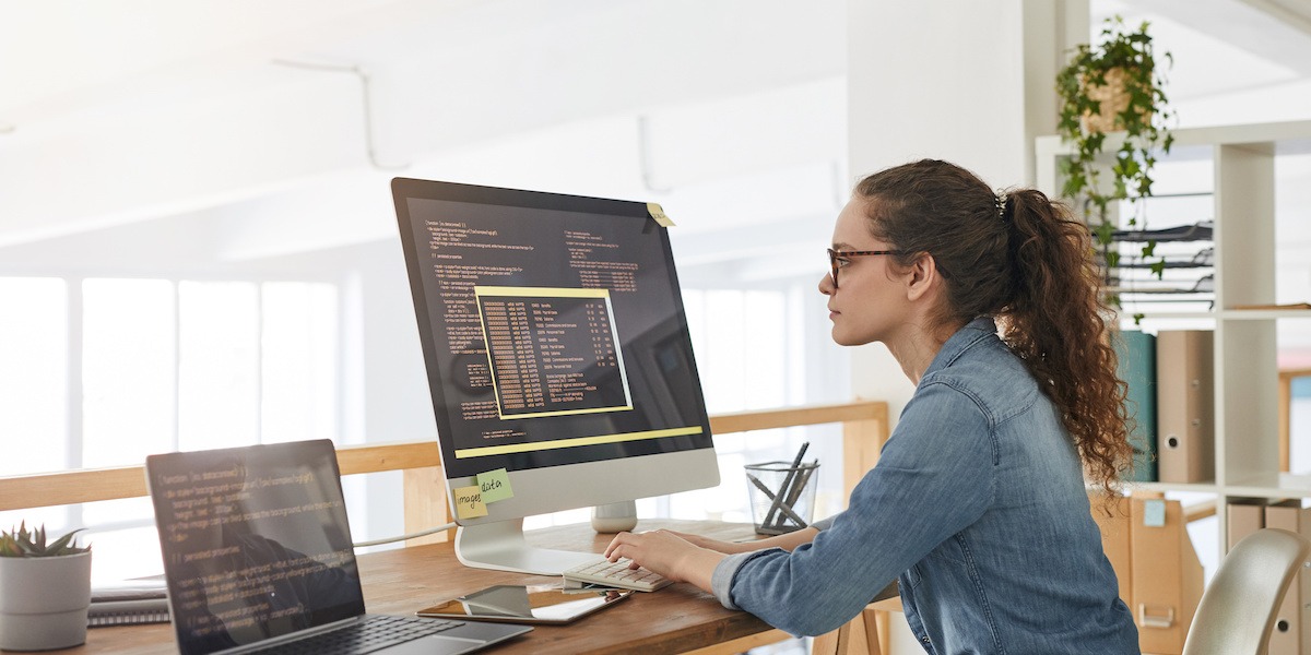 A software engineer sits in an office working on a desktop and a laptop screen.