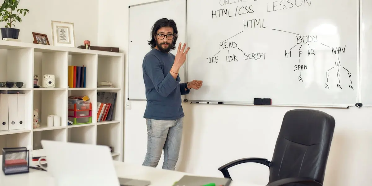 A teacher of an online coding class stands by a whiteboard facing his laptop teaching HTML and CSS.