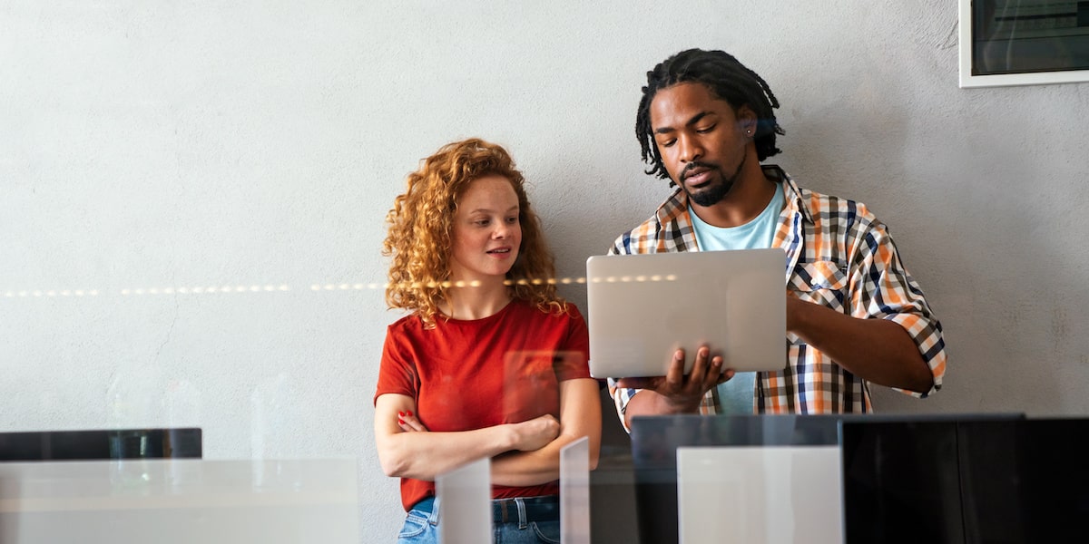 A programmer and a coder stand with a laptop in a startup office discussing something.