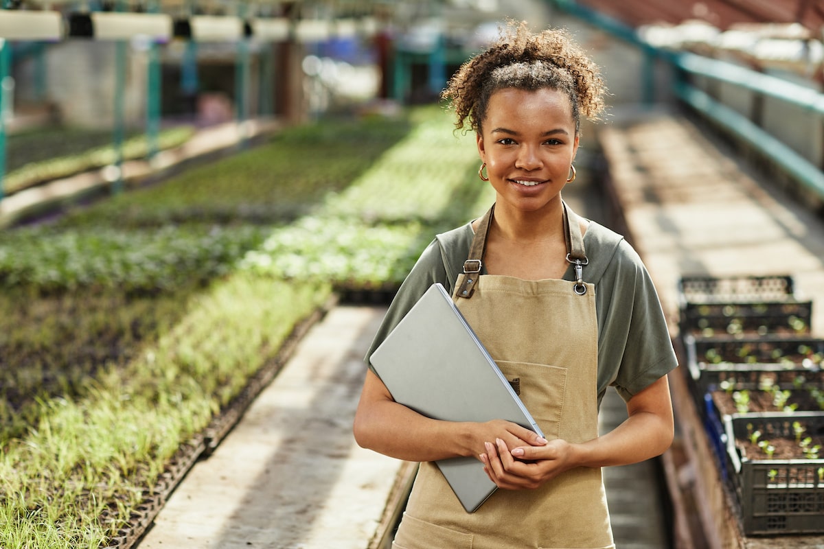 A woman stands with a laptop in a garden centre, wearing an apron.