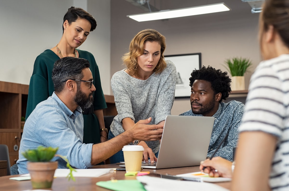 A product manager sits in a meeting in an office pointing out something on a laptop to their team.