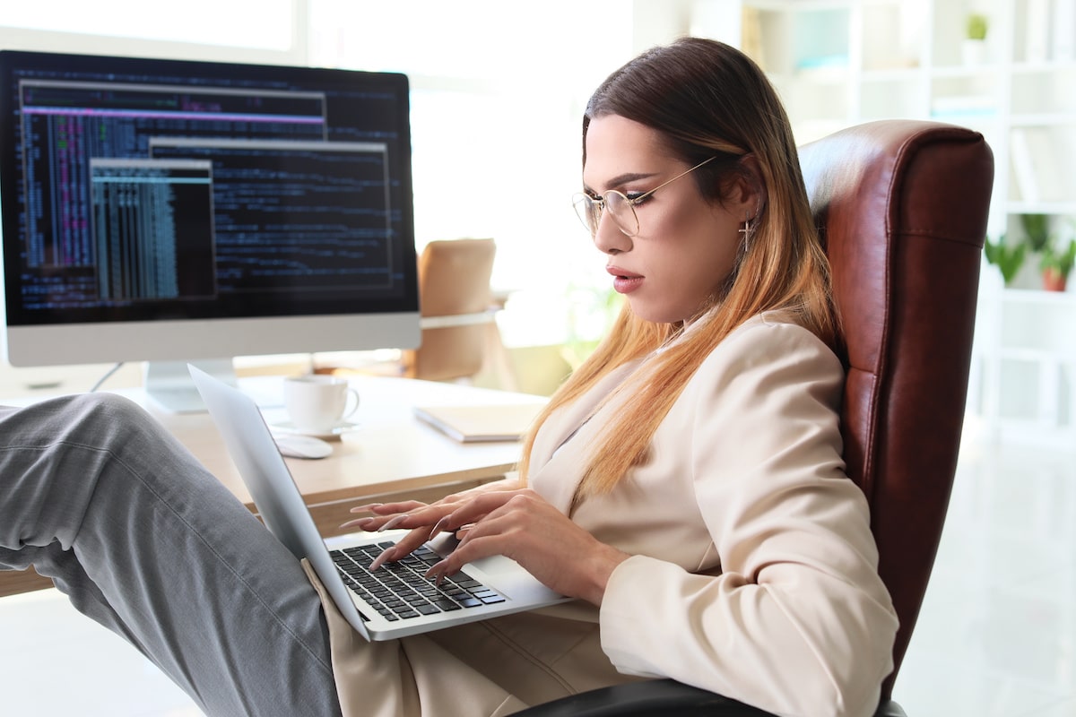 An entry-level programmer sits at her desk on her laptop, with code displaying on her monitor screen.