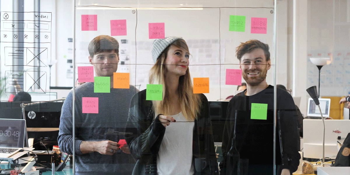 Students standing behind a glass wall covered in sticky notes, working on their UX design course
