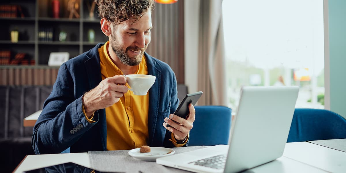 remote tech job a young man with a cup of coffee in his hand at his laptop looks at remote tech jobs on his cellphone
