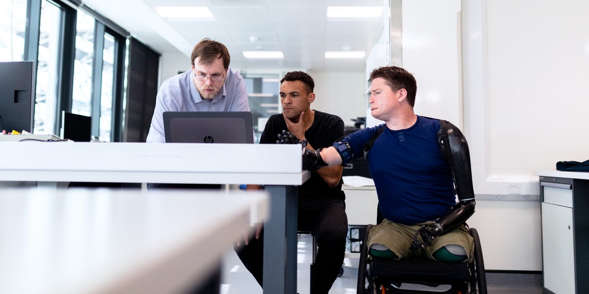 Three men sit around a screen, one in a wheelchair, to learn web development.
