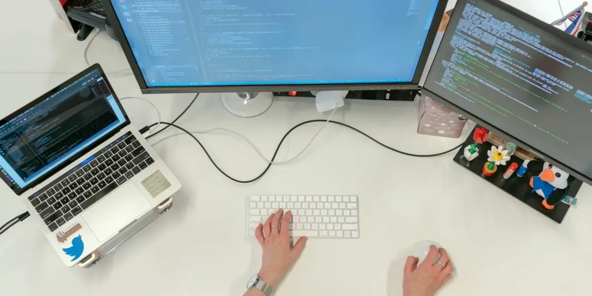 A web developer sits at two computer screens and a laptop, working with the Flask microframework