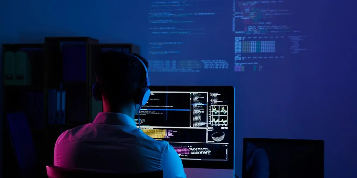 A Node.js developer sits at a desk coding on a computer, with more JavaScript code projected on the wall behind him.