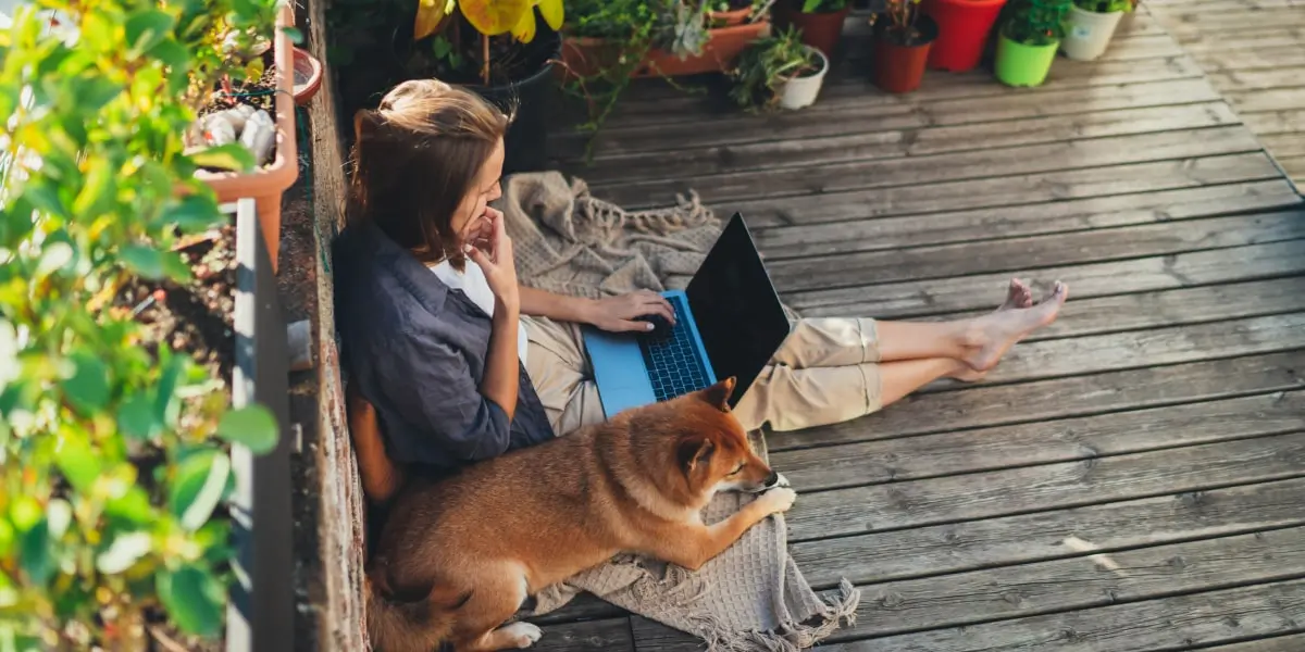 A coder working remotely sitting outside working on her frontend developer skills with her laptop and her dog beside her.