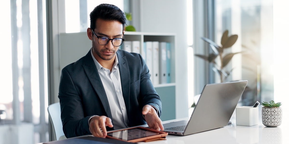 A young man with glasses reviews his tablet at his desk.