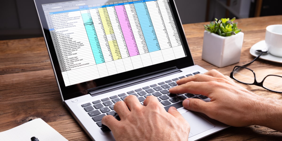 A pair of hands typing on a laptop keyboard, with a spreadsheet of structured data on the screen