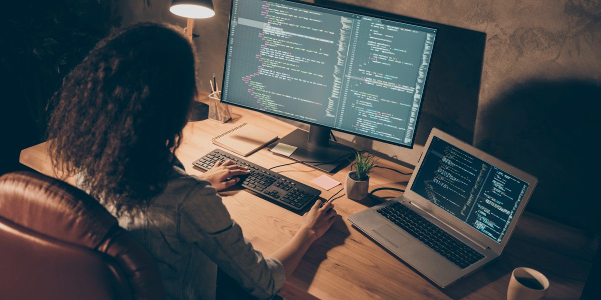 A web developer sitting at a desk, looking at code on two computer screens