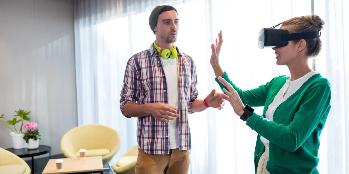 A woman in a VR headset stands beside a man assisting her, in an office.