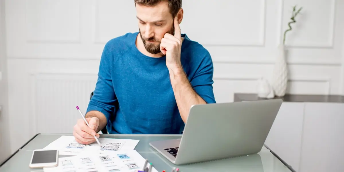 A UX designer sits at a desk, working at a laptop and paper with coloured pencils.