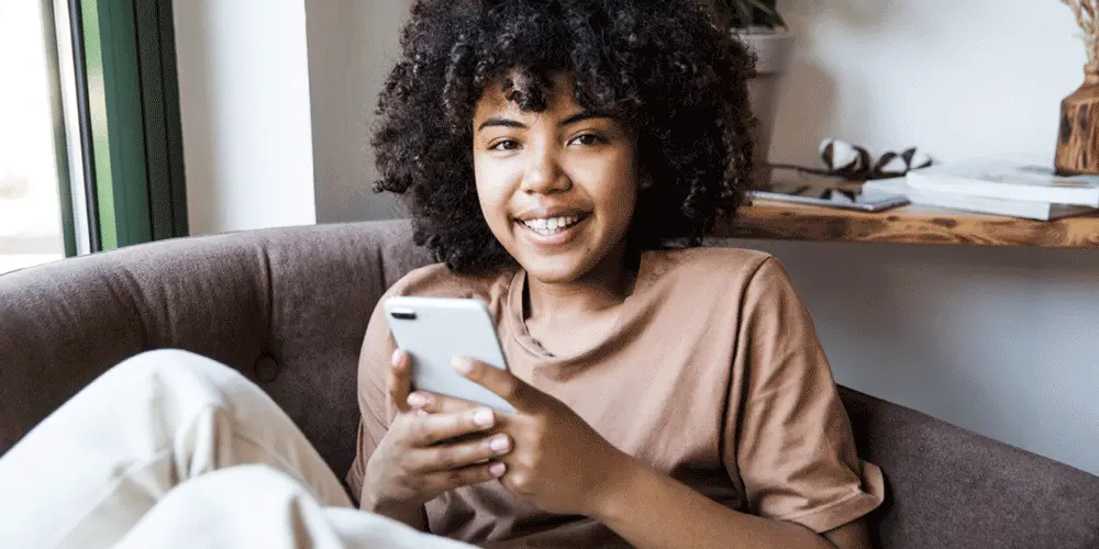 A person sitting on a couch, holding their mobile phone and smiling at the camera