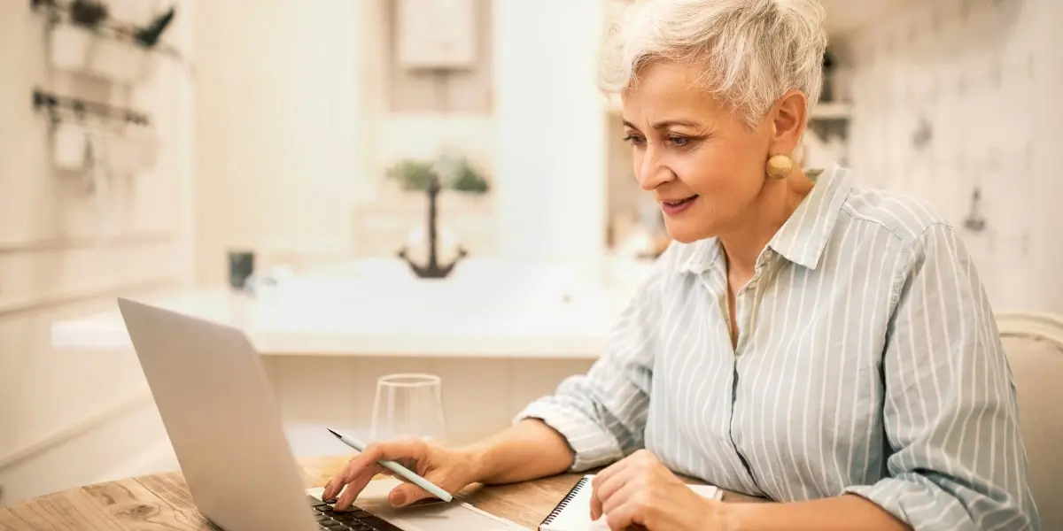 A grey-haired woman sits at her laptop learning to code at home on her laptop.