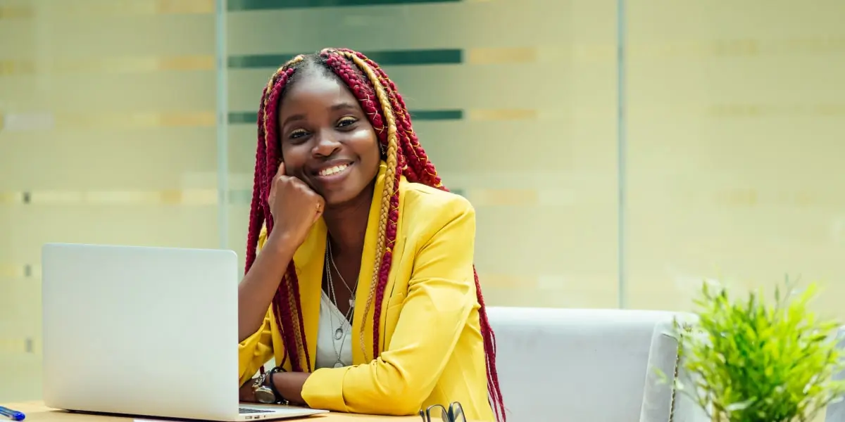 A smiling web developer sits at her desk in a startup office beside her laptop.