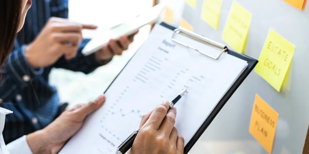 Two designers' hands, one pointing to a whiteboard, the other going through notes on a clipboard
