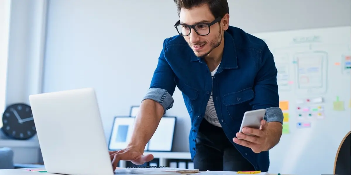 A UX designer standing at a desk, looking at a computer screen and holding a mobile phone