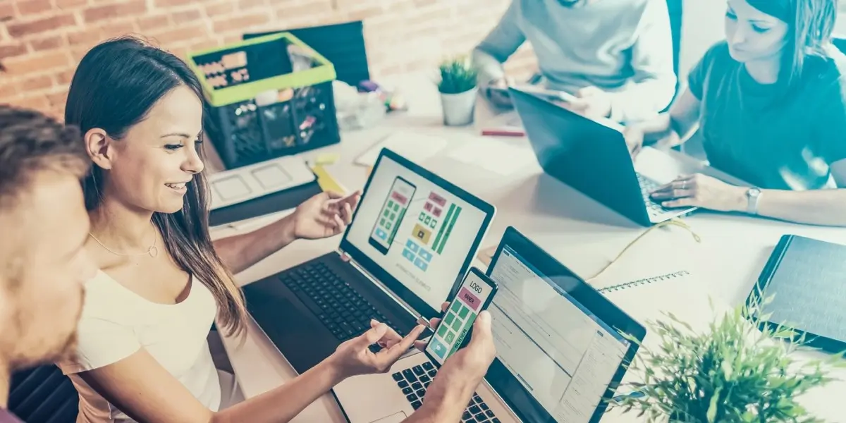 Designers sitting at a shared desk, working on their laptops and phones