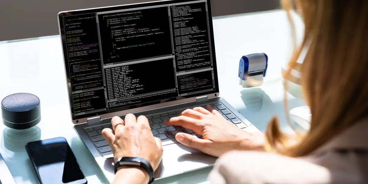 A web developer works away at a laptop screen full of code at her desk, with her phone, bluetooth speaker and stamp beside it.