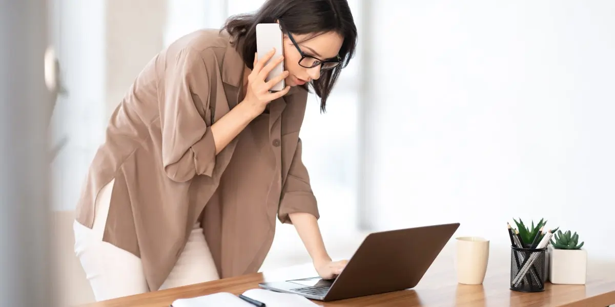 A startup worker looking at a laptop, speaking into a smartphone