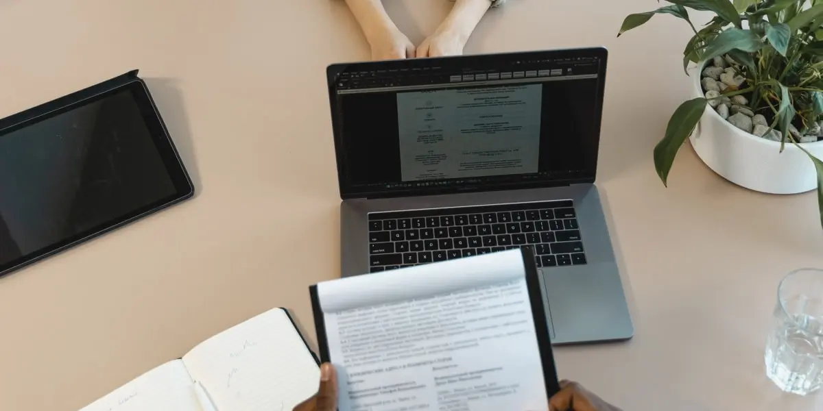 An interviewer holds a notepad and has a laptop and tablet on the table in front of them during an iOS developer interview