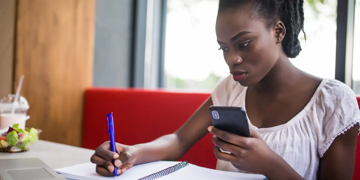 A digital marketing manager writing something in a notebook and holding a smartphone
