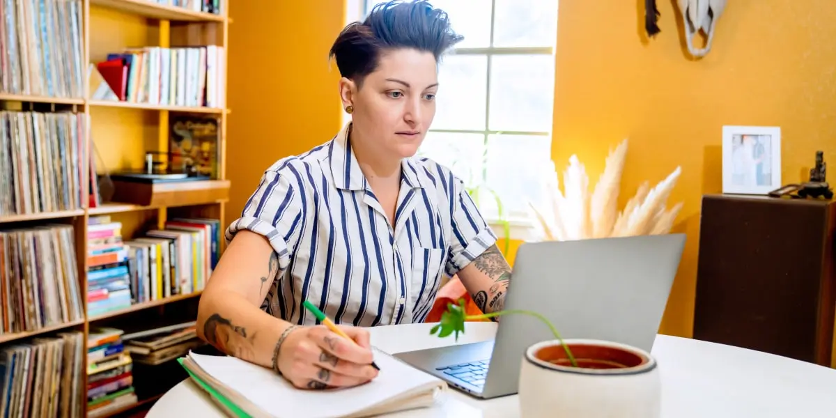 A digital marketing analyst working from home, looking at a laptop screen and writing notes in a book