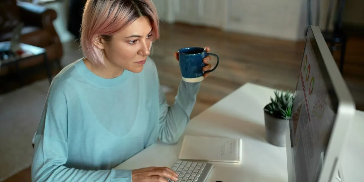 A web developer sits at her desktop PC drinking coffee and learning about CSS preprocessors on the screen.