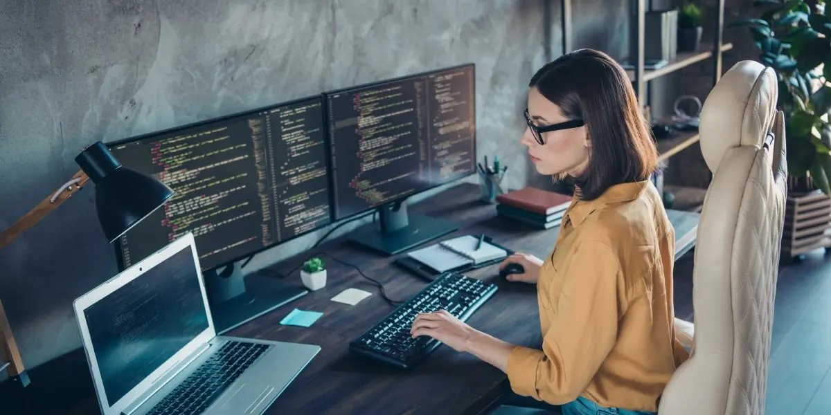 A software engineer sits coding in front of 3 monitor screens and a laptop.
