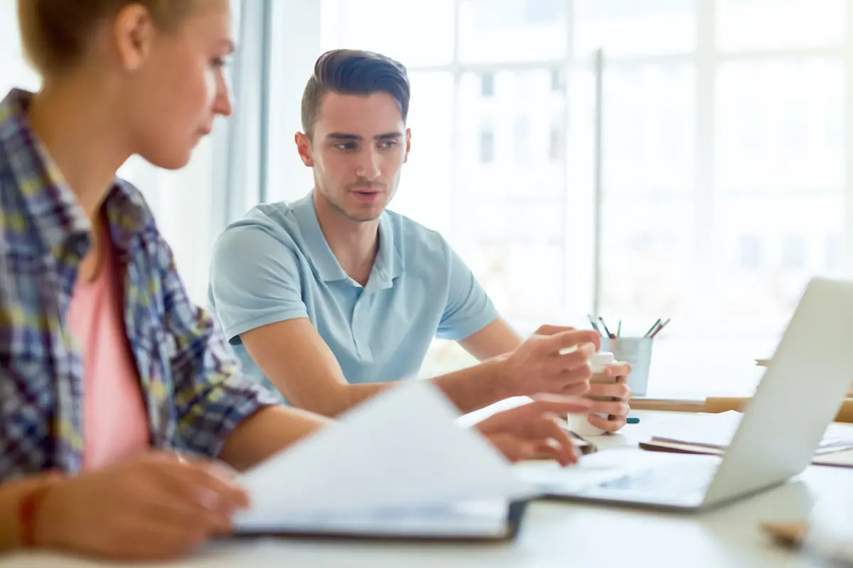 A woman in an office wondering what is a scrum master has it explained to her by her colleague.