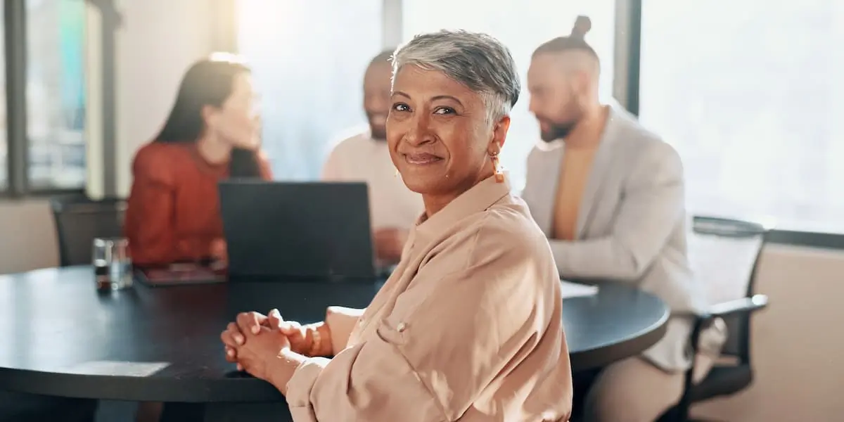 A scrum master leading her team faces the camera with a smile while they continue to work at a desk