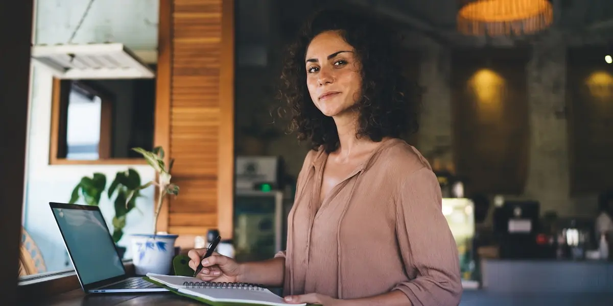 A remote worker stands at a desk with her laptop and a notepad