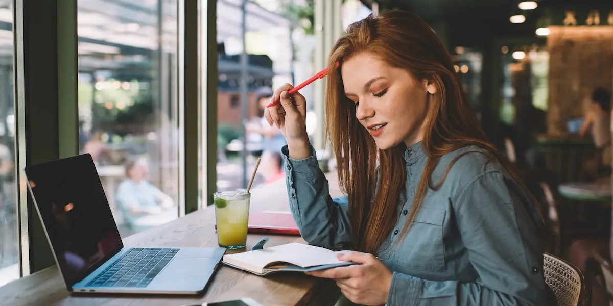 product design courses A young redhead sits in a coffee shop at the window with a notepad and computer looking up the best product design courses