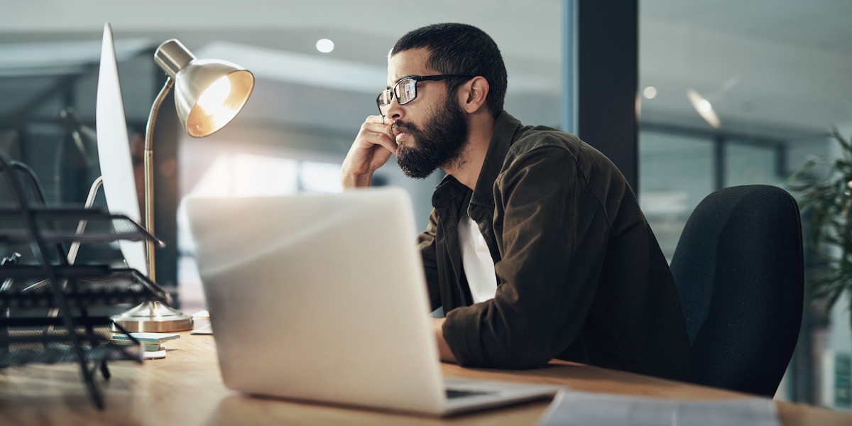 A young bearded man with glasses sits at a desktop computer with laptop open researching how to become a product designer