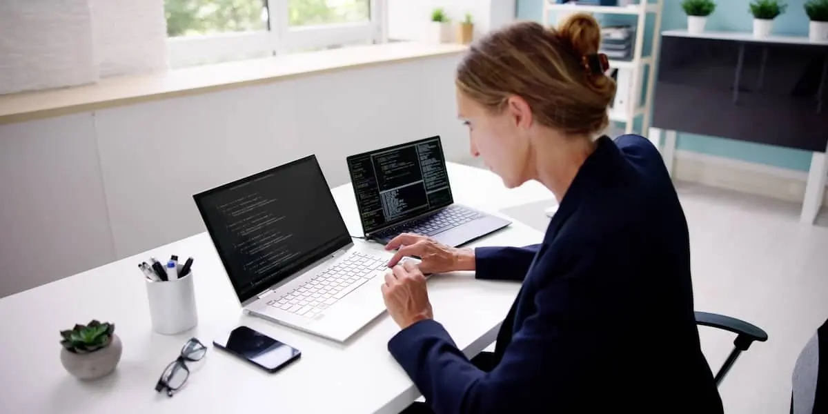 An analytics engineer sits in a bright home office with two laptops of code.