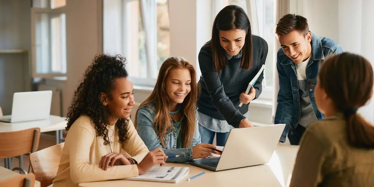 A group of high school students huddles around a laptop, smiling.