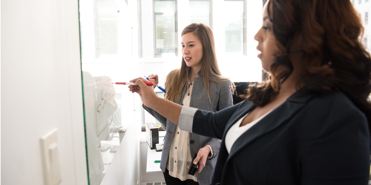 Two women standing at a whiteboard, writing and discussing ideas