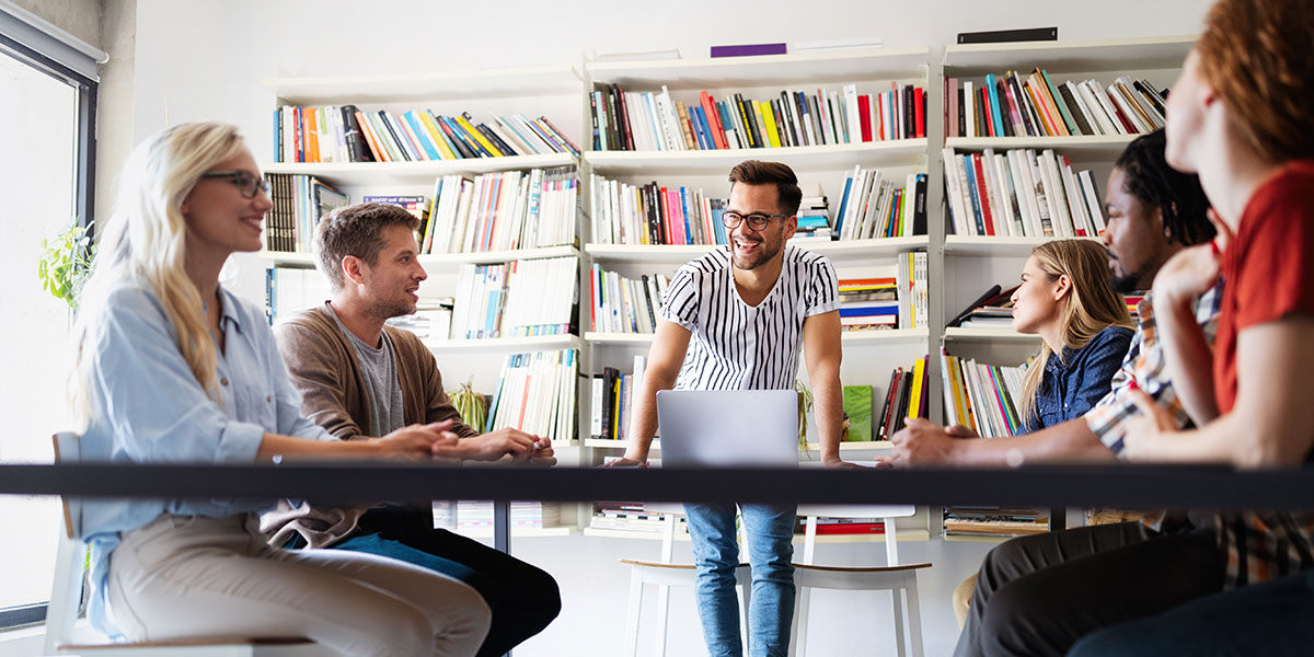 A designer standing up in front of a group of people sat around a table A UX designer stands at a desk leading five seated designers in the ideation design process