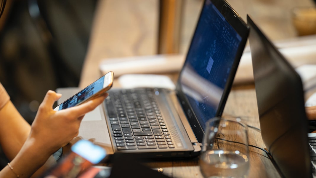 A user sitting at a table in an open-plan office, working at a laptop and looking at their phone