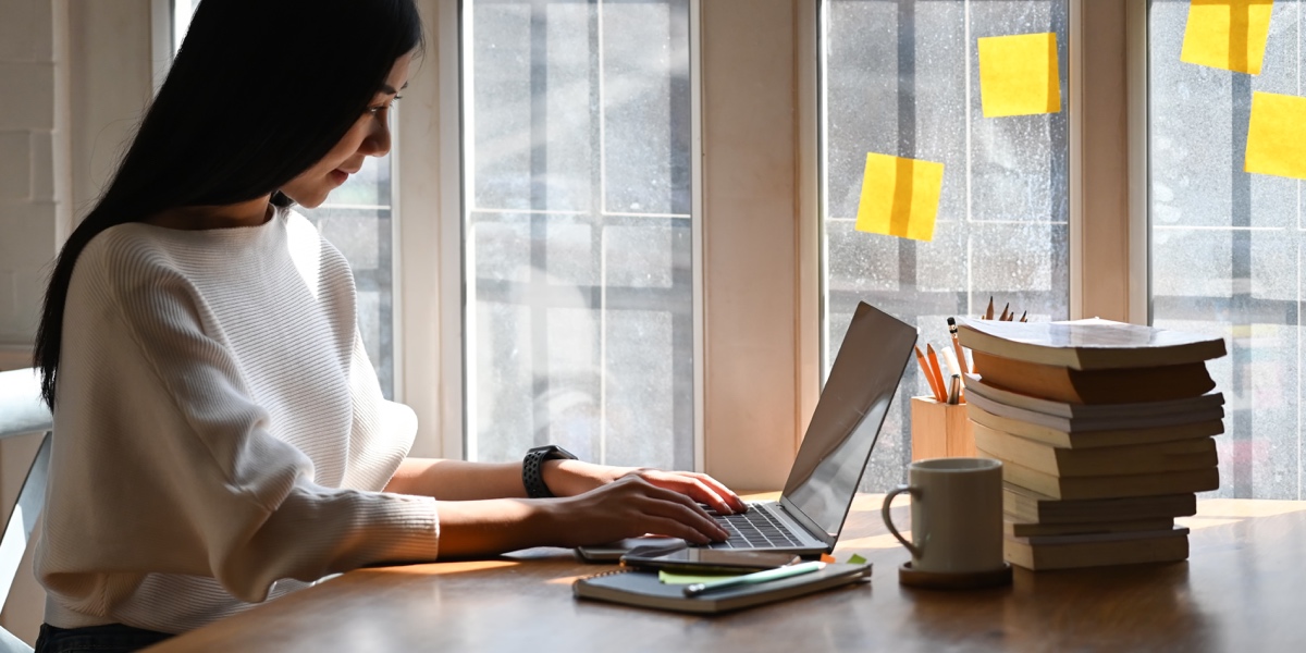 The Power of Storytelling in UX A designer working at a laptop on a desk with a stack of books, near a sunny window covered in sticky notes