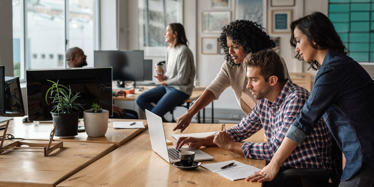 Three designers gathered around a desk discussing a project with a cup of coffee and a laptop on the desk, two colleagues conversing in the background