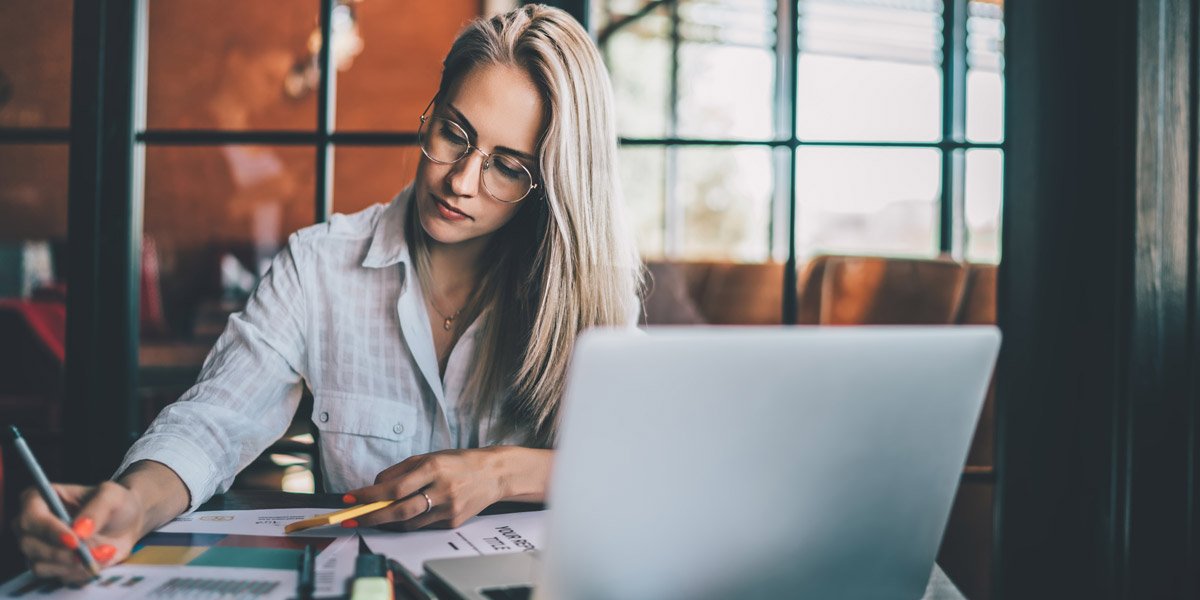 A UX design student sitting at her desk, behind a laptop, taking notes with paper and pen